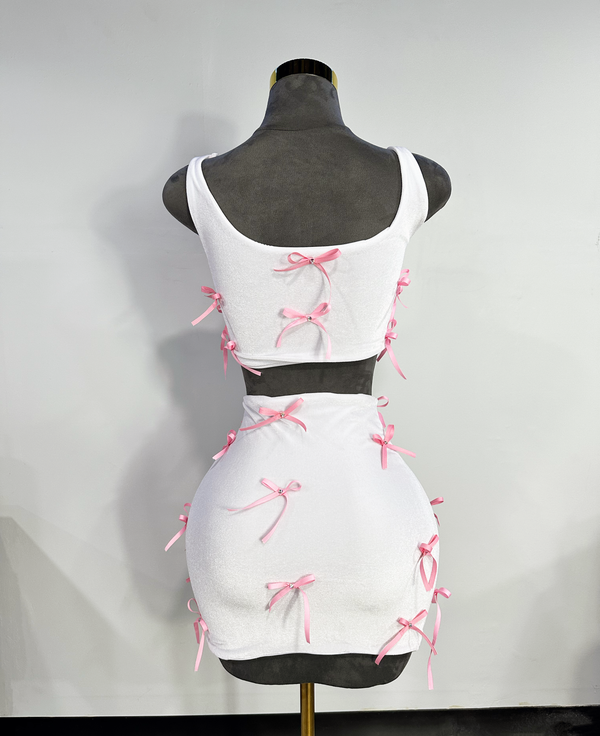 A two-piece skirt set with a white fitted tank top and a white high-waisted mini skirt, both adorned with pink ribbon bows and rhinestone accents by Bellz Couture in Detroit. shown on mannequin against a plain wall.