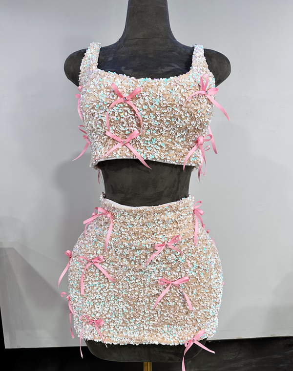 A two-piece outfit with a sequined tank top and a matching mini skirt, both adorned with pink ribbon bows and rhinestone accents by Bellz Couture of Detroitdisplayed on a mannequin.