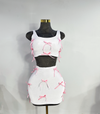 A two-piece skirt set with a white fitted tank top and a white high-waisted mini skirt, both adorned with pink ribbon bows and rhinestone accents by Bellz Couture in Detroit. shown on mannequin against a plain wall.